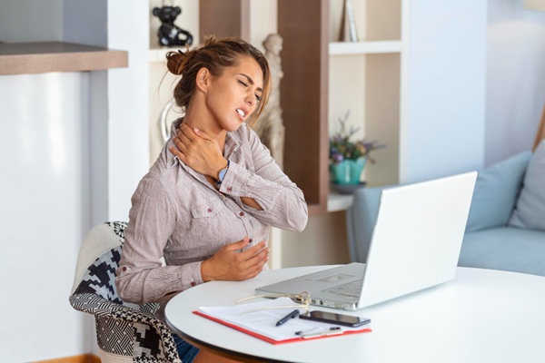 woman-dealing-with-neck-strain-at-desk Woman sitting at a desk holding her neck due to muscle strain and posture-related tension while working on a computer